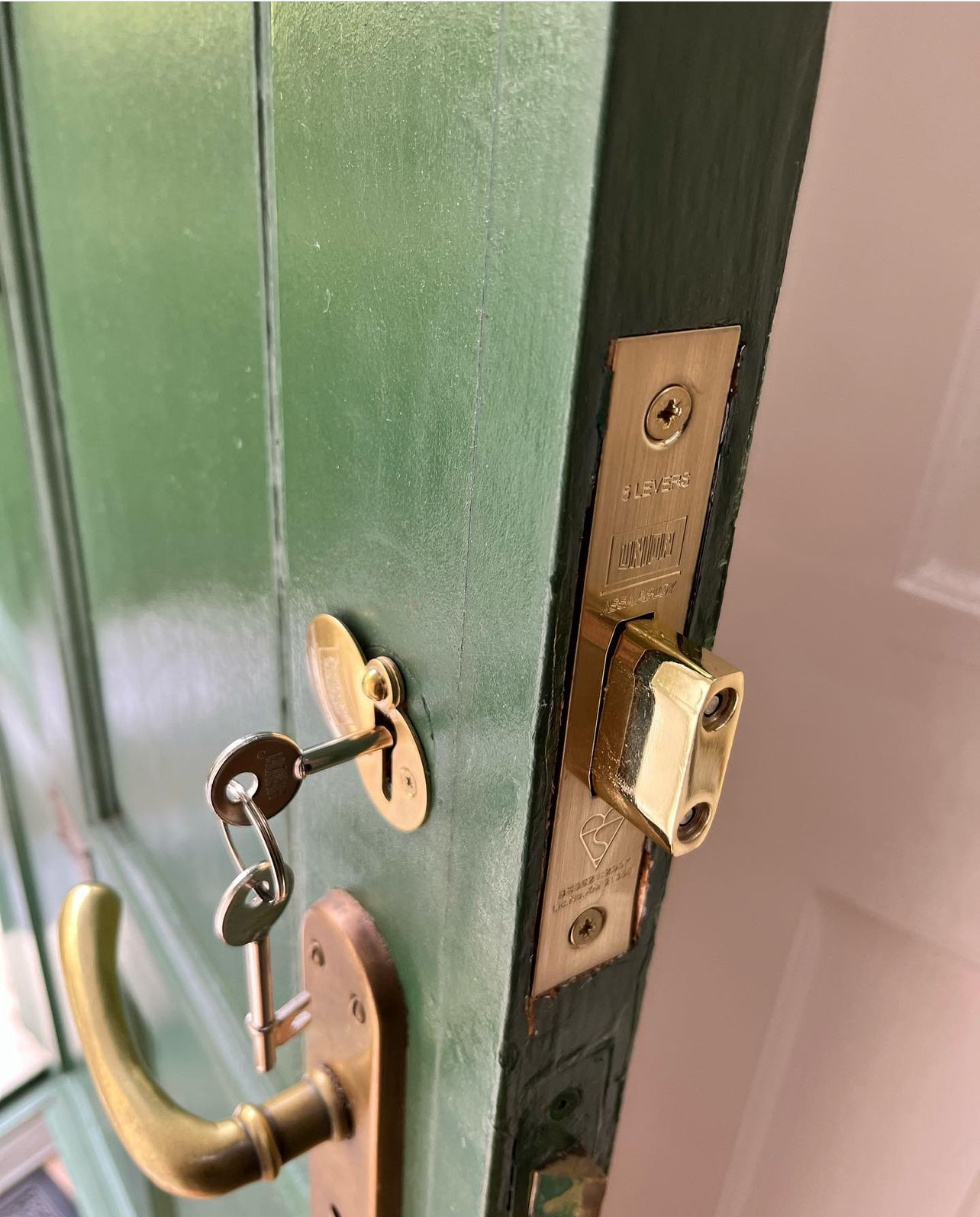 Close-up of a green wooden door with a brass mortice lock and traditional lever key inserted, showing the lock mechanism and handle.