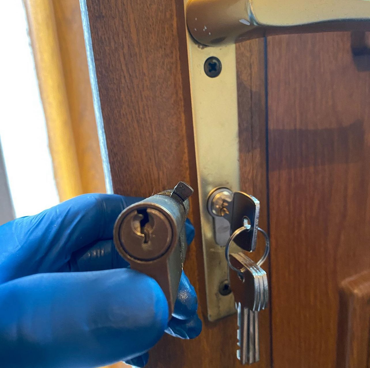 Close-up of a locksmith’s hand wearing blue gloves replacing a Euro cylinder lock in a wooden door with keys hanging from the lock.